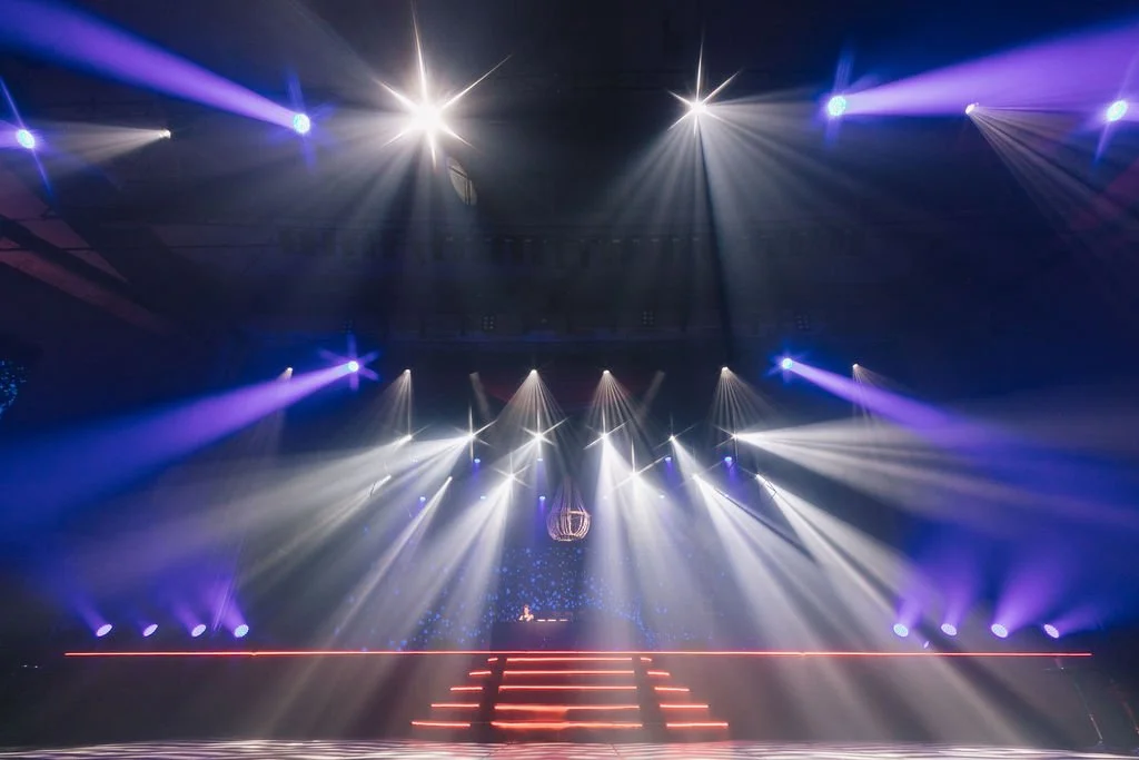 Palco de evento com luzes de palco em tons de branco, azul e roxo, com degraus iluminados em vermelho.