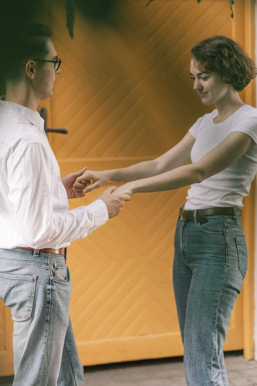 Duas pessoas, uma de camiseta branca e calça jeans, e outra de camisa branca com calça jeans, estão se segurando pelas mãos em um ambiente fechado com parede de madeira ao fundo.
