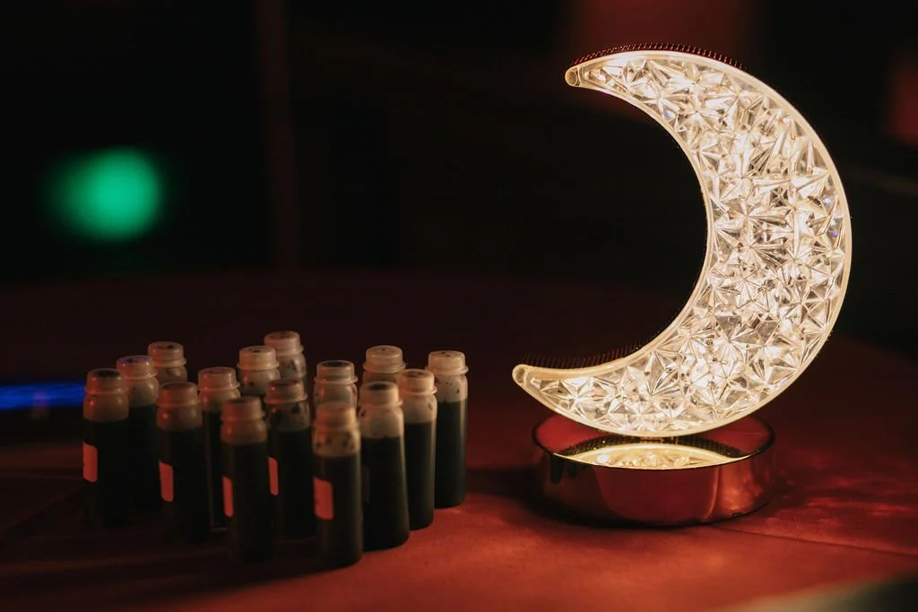 Luzes de uma luminária de formato de lua crescente ao lado de várias ampolas de substância escura sobre uma mesa de madeira.