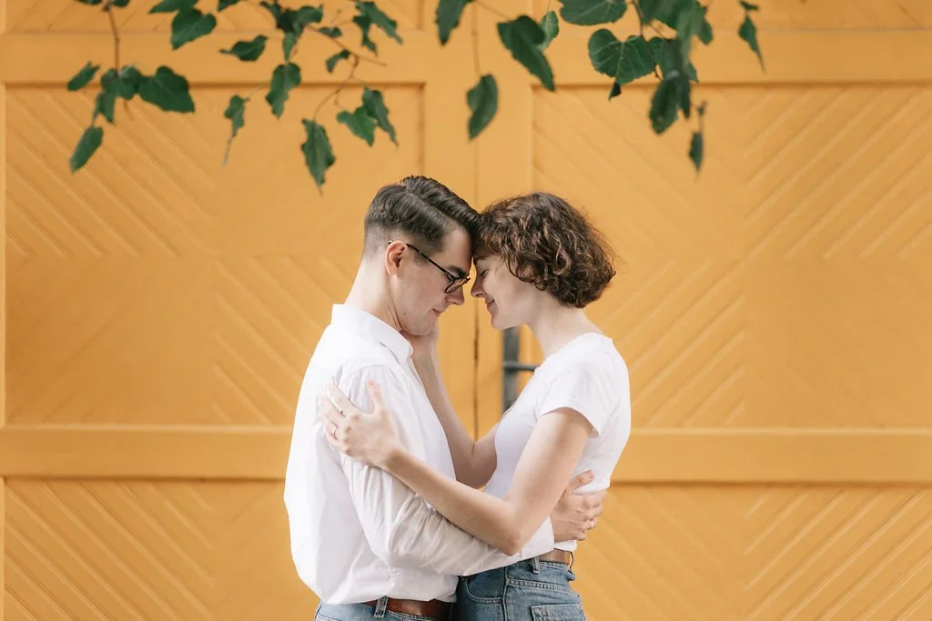 Casal jovem se abraçando, com a testa unida, diante de uma parede de madeira clara decorada com folhas verdes.