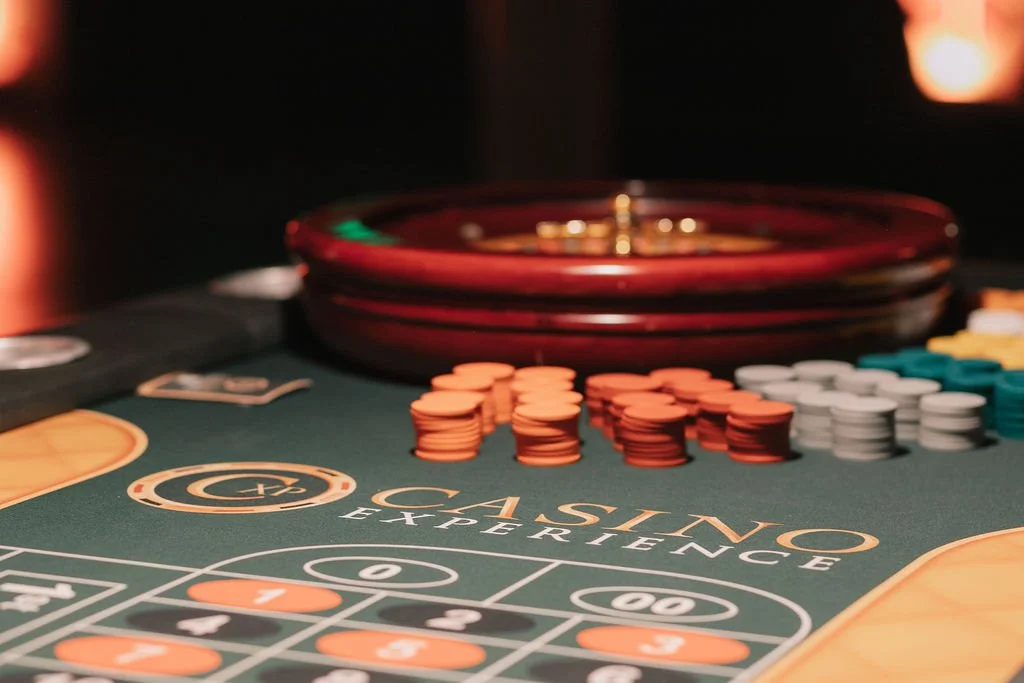 Mesa de blackjack com fichas de apostas e uma roleta ao fundo.