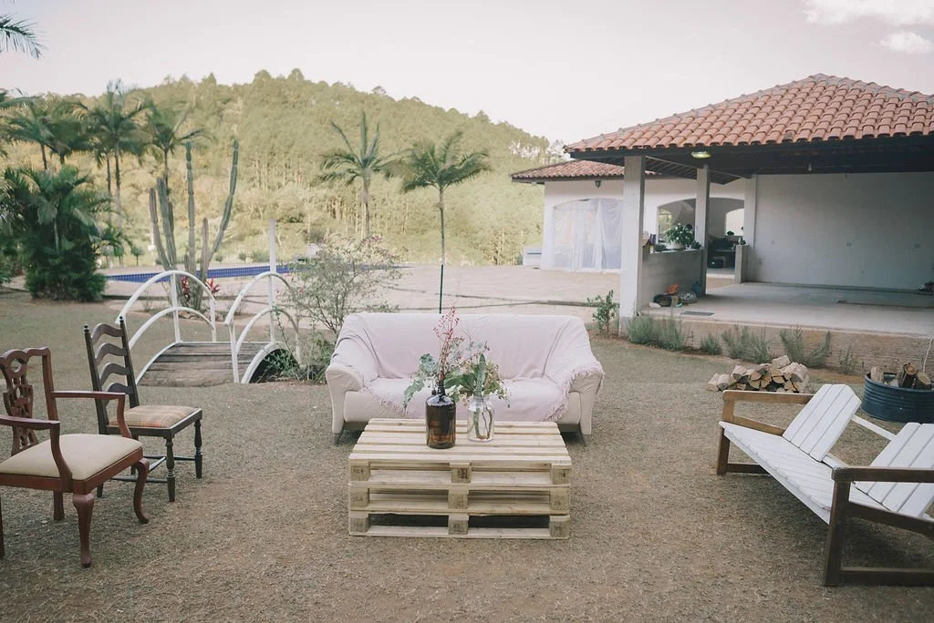 Área ao ar livre com sofá de almofada, cadeiras de madeira, mesa de pallets, jardim, árvores, casa com estrutura coberta e esquecido de fogo ao lado.