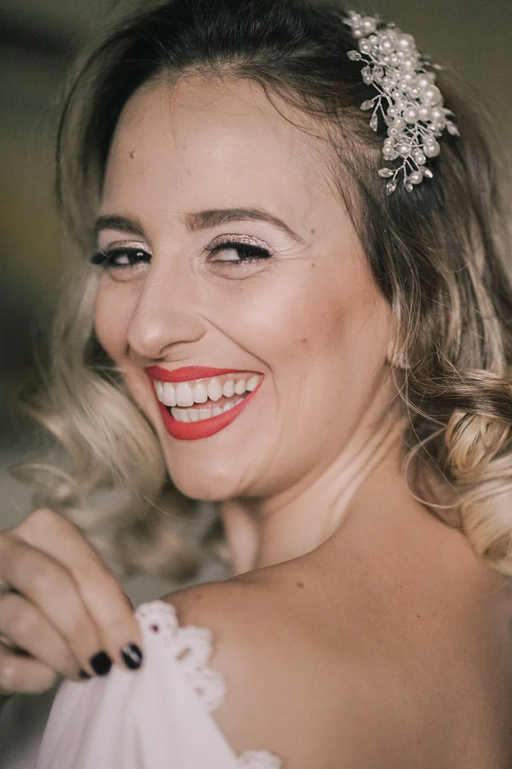 Mulher sorridente com maquiagem, cabelo cacheado e acessório de casamento com pérolas na cabeça.