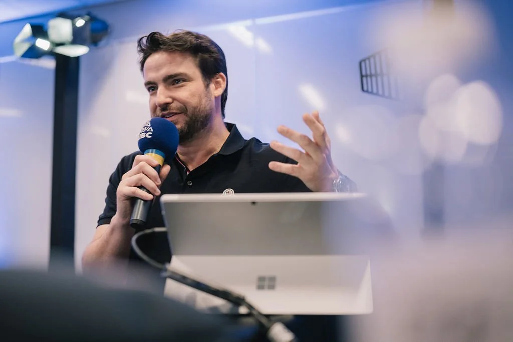 Homem falando em um microfone durante uma apresentação ou palestra com fundo desfocado.