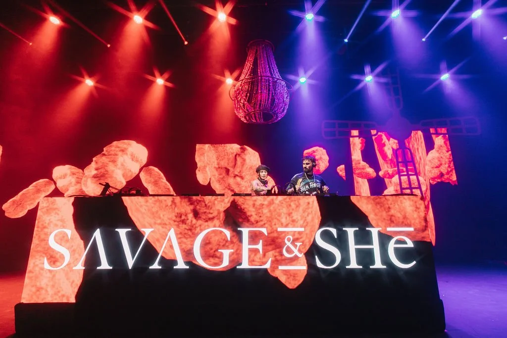 DJs tocando em um palco com iluminação colorida, tela de fundo com imagens de rochas, e o nome 'SAVAGE & SHE' exibido.