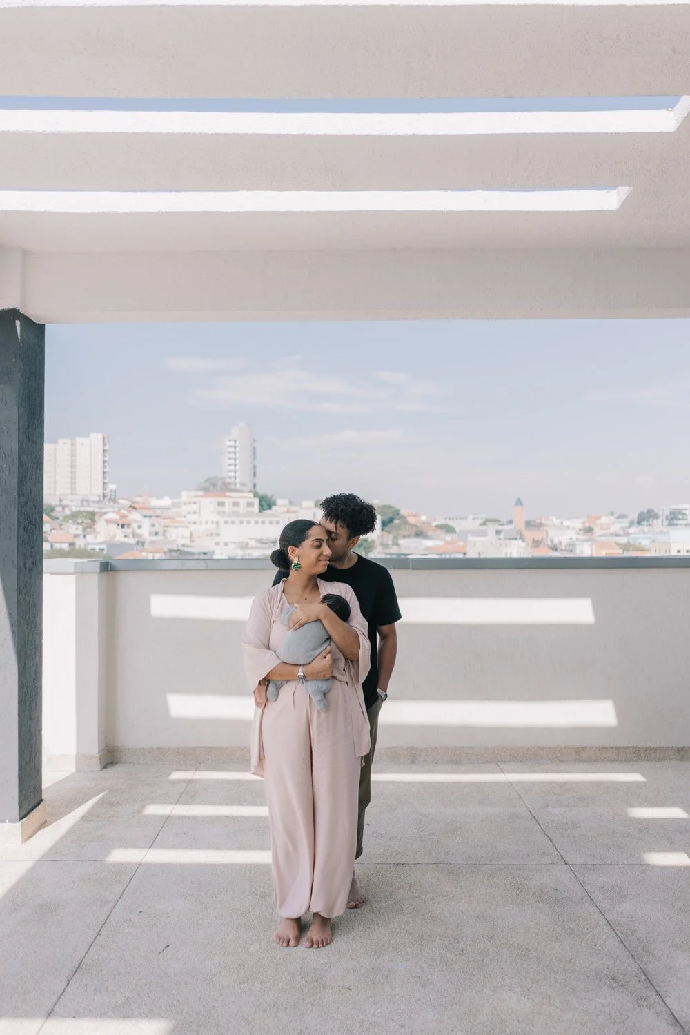 Família com bebê no terraço de um prédio, com vista da cidade ao fundo, dia ensolarado.