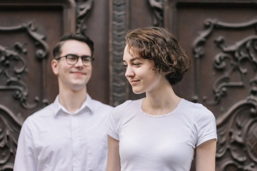 Um homem e uma mulher com roupas brancas, com mulher na frente e homem no fundo, diante de uma porta de madeira com detalhes decorativos, ambos sorrindo levemente.