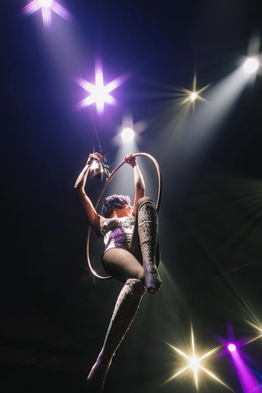 Artista de circo realizando apresentação com aye aérea sob luzes brilhantes no palco.