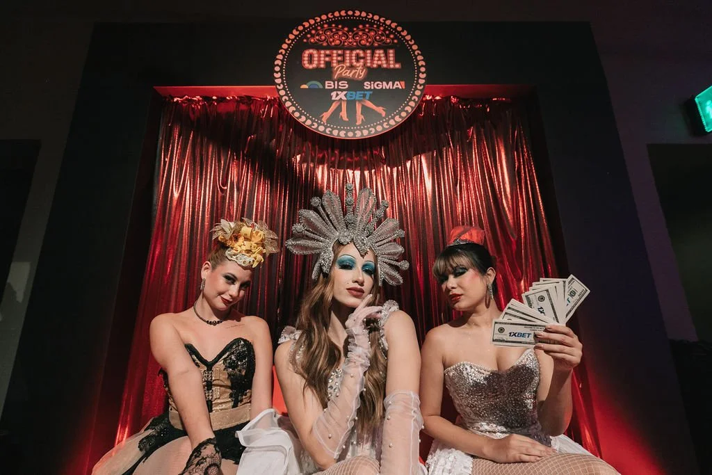 Três mulheres em trajes extravagantes e acessórios elaborados, sentadas em frente a cortinas de tecido vermelho, em um cenário de festa ou evento, com um letreiro que diz 'Oficial Party' no fundo, acima de um logo de patrocinadores.