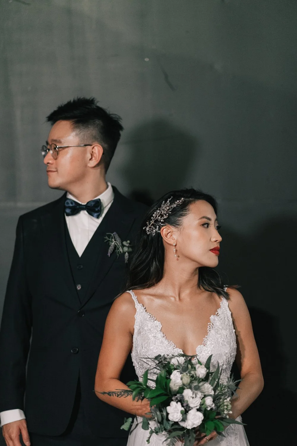 Casal de noivos em traje de casamento, a noiva segura um buquê de flores brancas e verdes. Ele usa um terno preto com gravata borboleta, ela usa vestido de noiva branco com detalhes de renda e acessórios de cabelo com brilho.