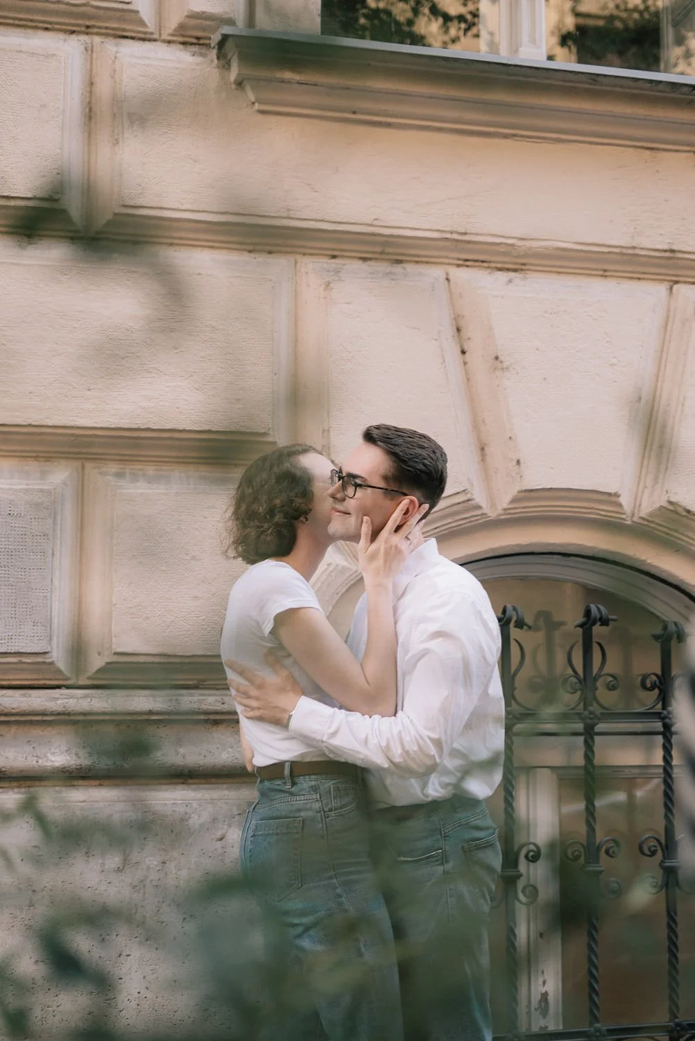 Casal apaixonado se abraçando e se beijando na rua, com fachada de prédio antigo ao fundo e vegetação ao redor.