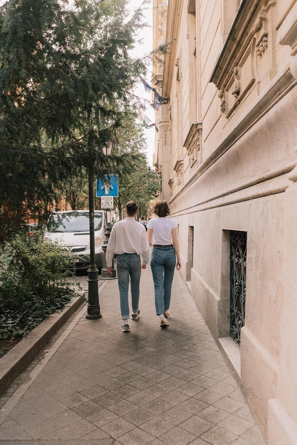 Casal caminhando de mãos dadas por uma calçada urbana, com árvores e veículos estacionados ao lado, ao lado de um edifício antigo de paredes de pedra.