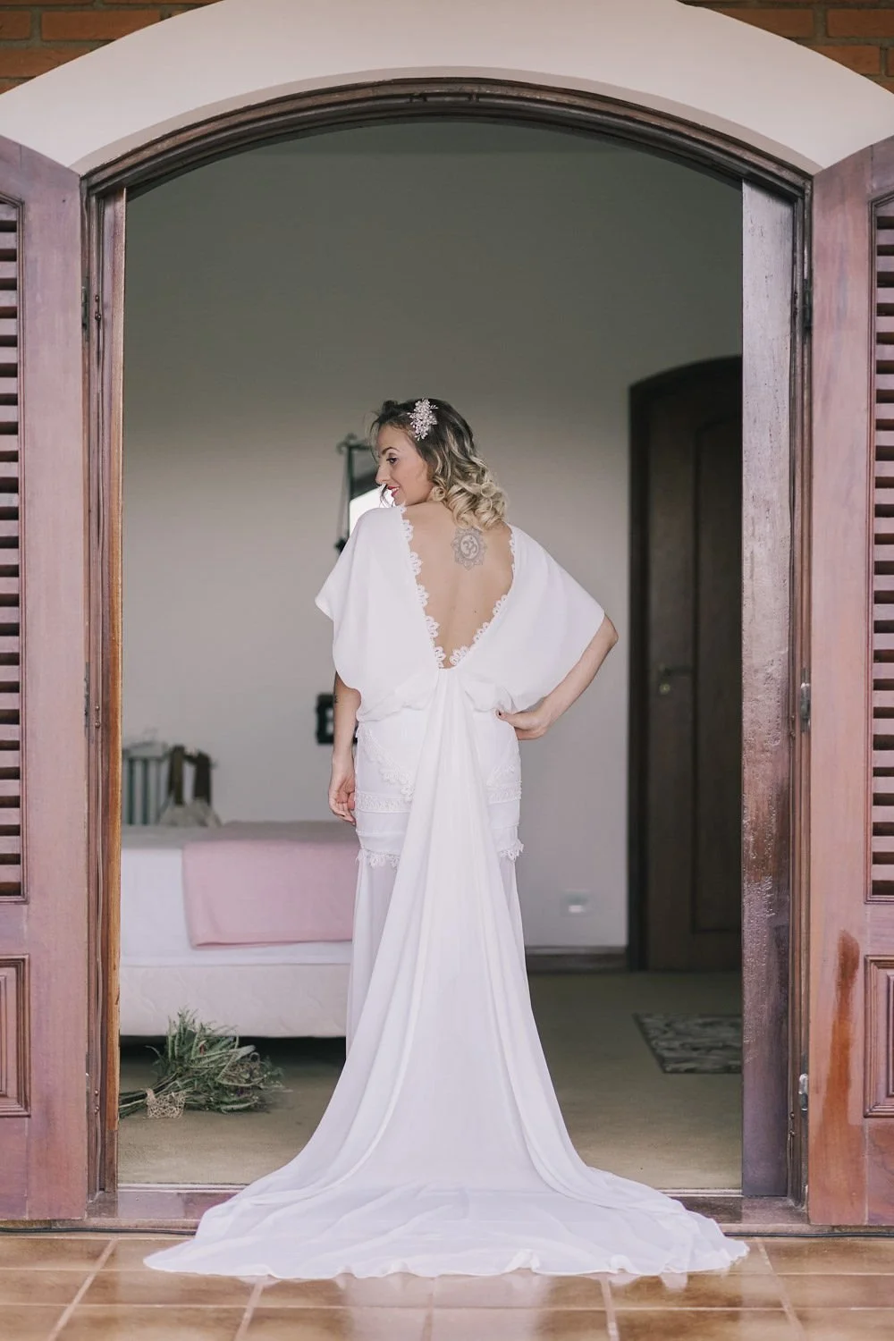Mulher de vestido branco com renda na parte de trás, de cabelo cacheado e enfeite na cabeça, posando na porta de uma casa