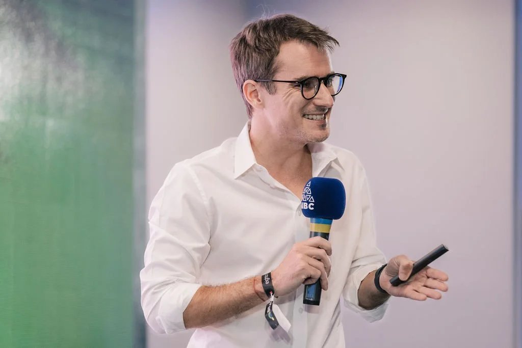 Homem sorridente segurando microfone e celular durante entrevista ou apresentação, vestindo camisa branca e óculos, ao lado de fundo cinza e verde.