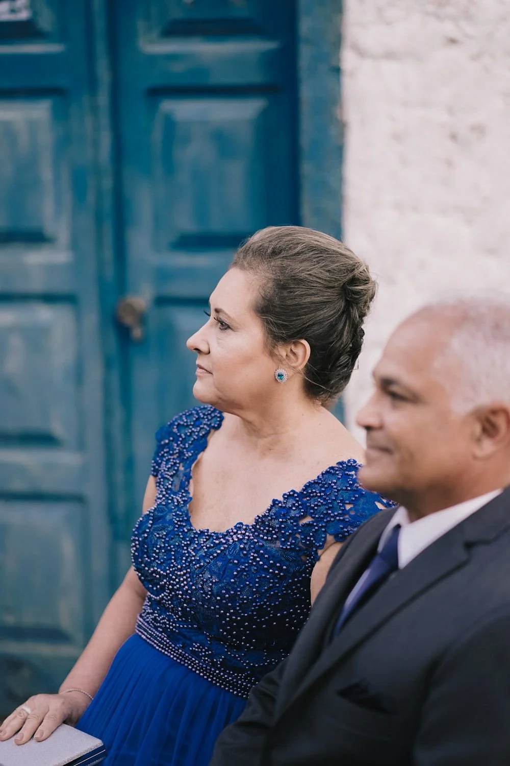 Mulher idosa, com vestido azul com detalhes de pedras, usando brincos de pedra azul, sentada ao lado de um homem de terno preto e gravata, em um ambiente com paredes brancas e uma porta azul ao fundo.