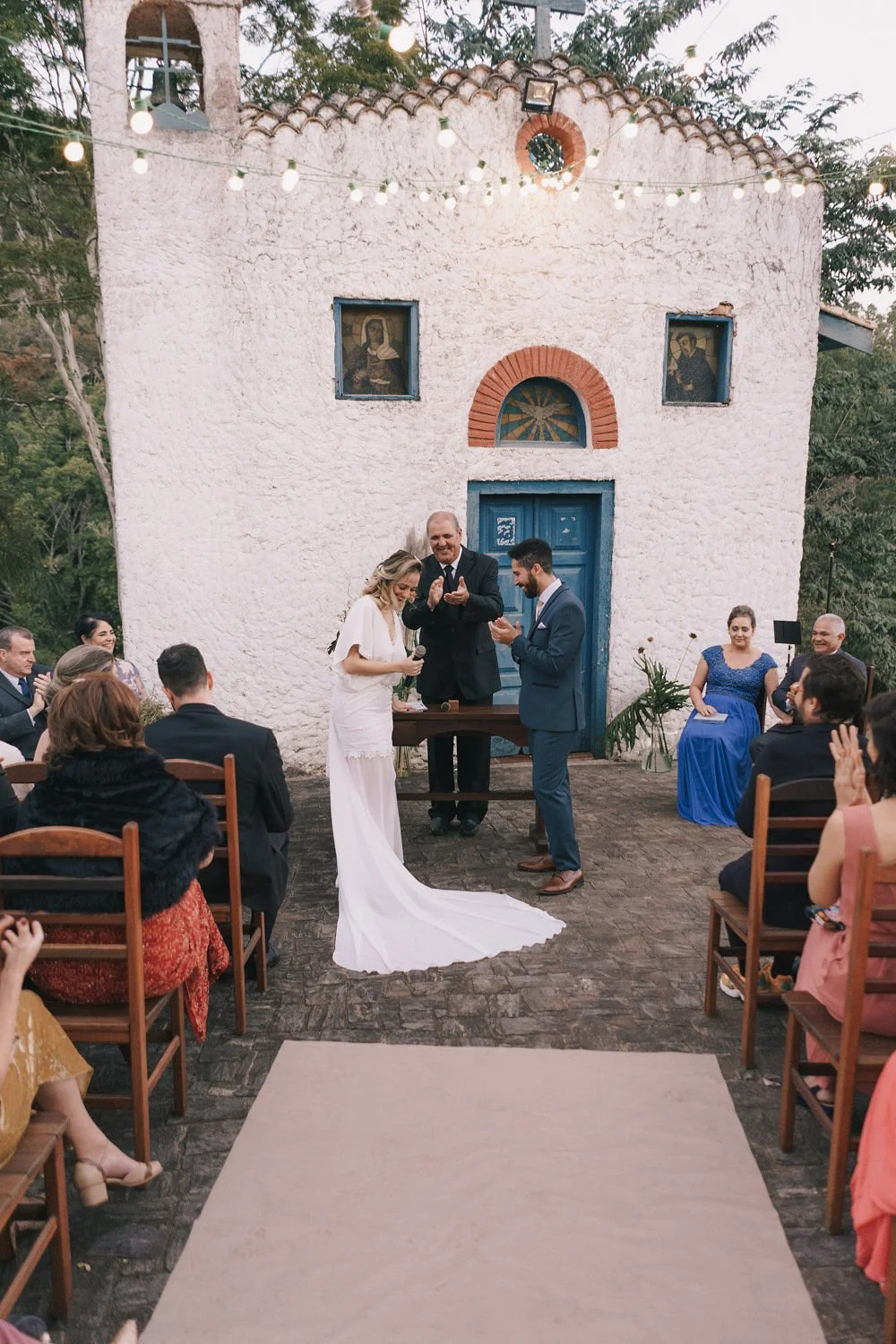 Casamento ao ar livre com os noivos no centro, cercados por convidados. O altar é em frente a uma pequena capela de parede branca, com teto de telhas e janelas com imagens religiosas. Decoração de luzes penduradas e plantas ao lado da porta. Os convi