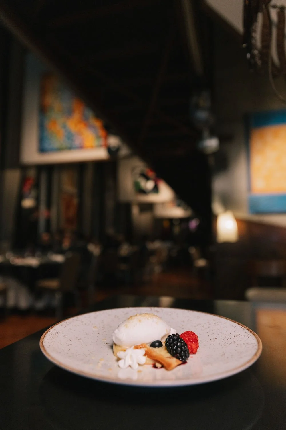 Sobremesa com sorvete e frutas, como amoras e framboesas, em um prato branco grande, em um ambiente de restaurante ou café decorado com quadros na parede.