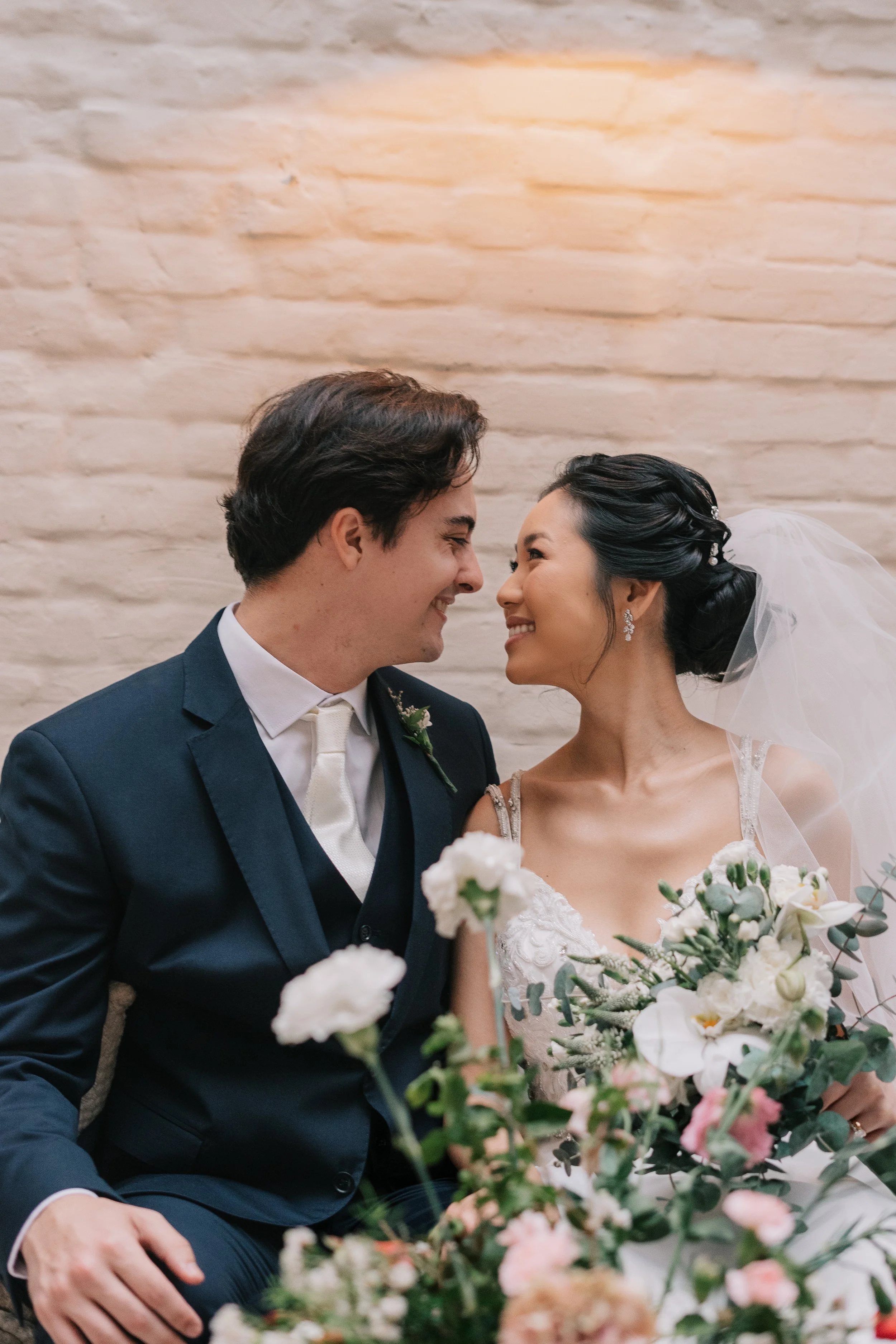 Casal de noivos sorrindo e se olhando com uma parede de tijolos ao fundo, a noiva segurando um buquê de flores brancas e verdes.