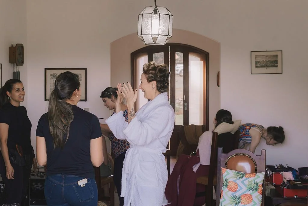 Mulheres conversando e rindo em um ambiente interno, com uma delas vestida de roupão branco, e duas outras na parede ao fundo, enquanto uma mulher se ajusta na cadeira.