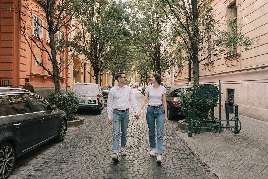 Casal jovem caminhando de mãos dadas na rua de paralelepípedos, com carros estacionados ao lado, árvores ao redor e edifícios antigos ao fundo.