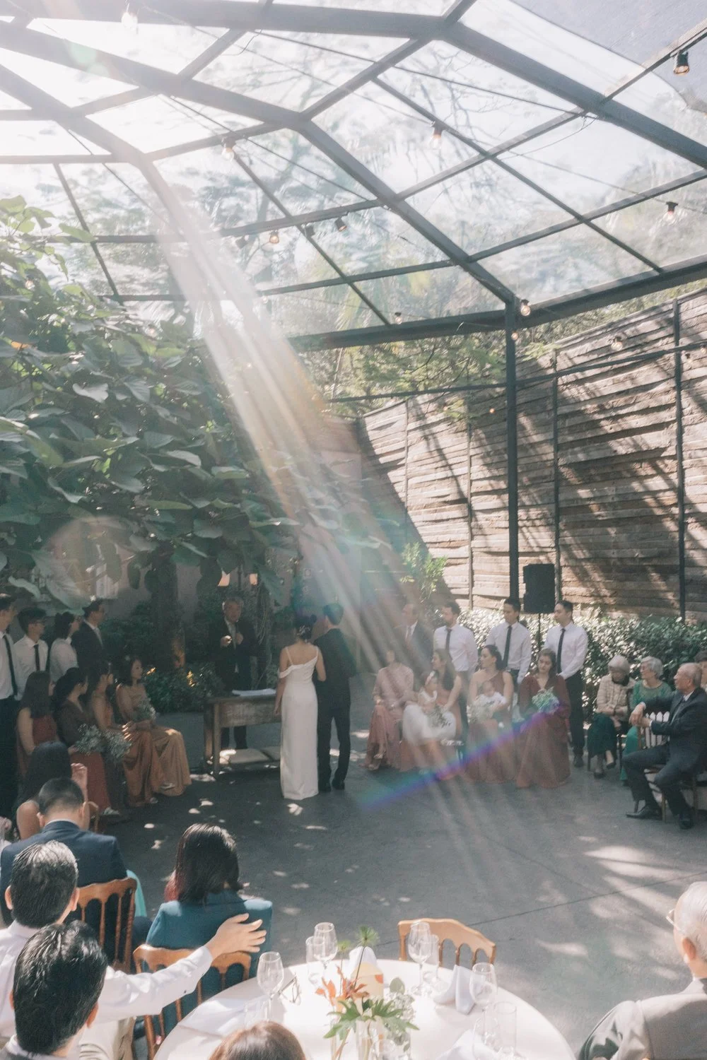 Cerimônia de casamento ao ar livre sob uma estrutura de vidro, com convidados sentados ao redor, alguns de pé, e o casal no centro trocando votos, com luz solar entrando por frestas.