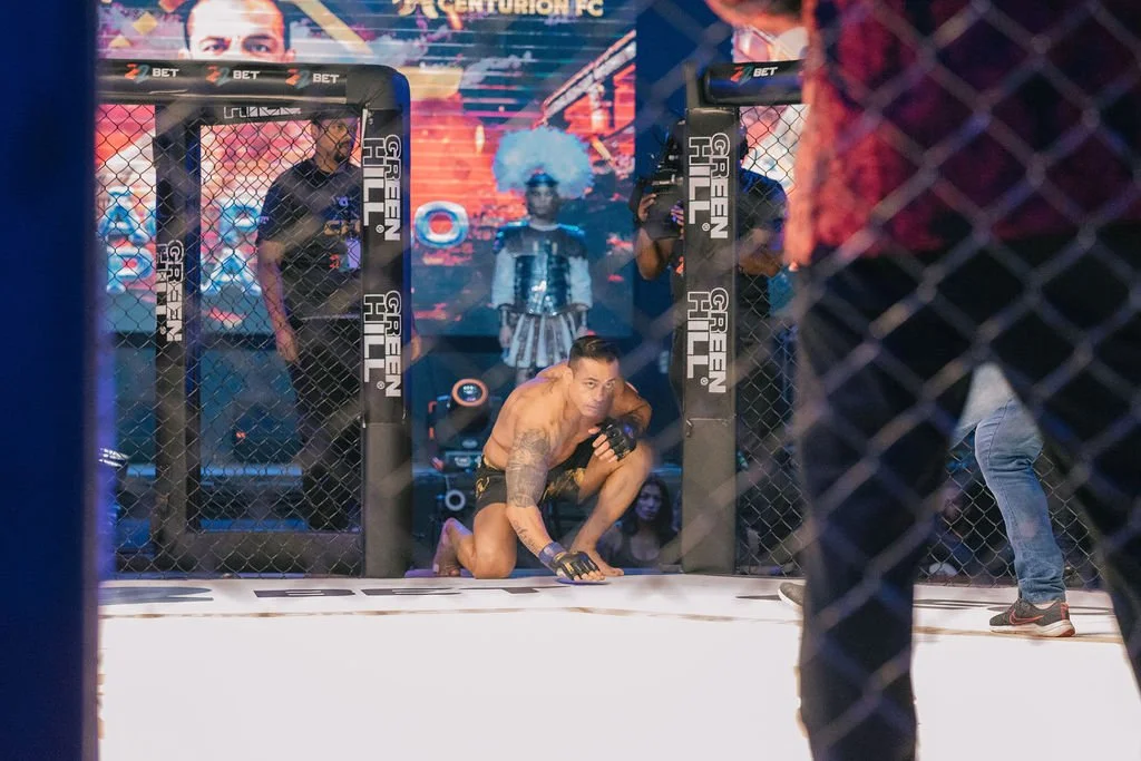 Lutador sentado no chão dentro de uma jaula de MMA, aparentemente após uma luta, rodeado por pessoas e equipe de apoio.
