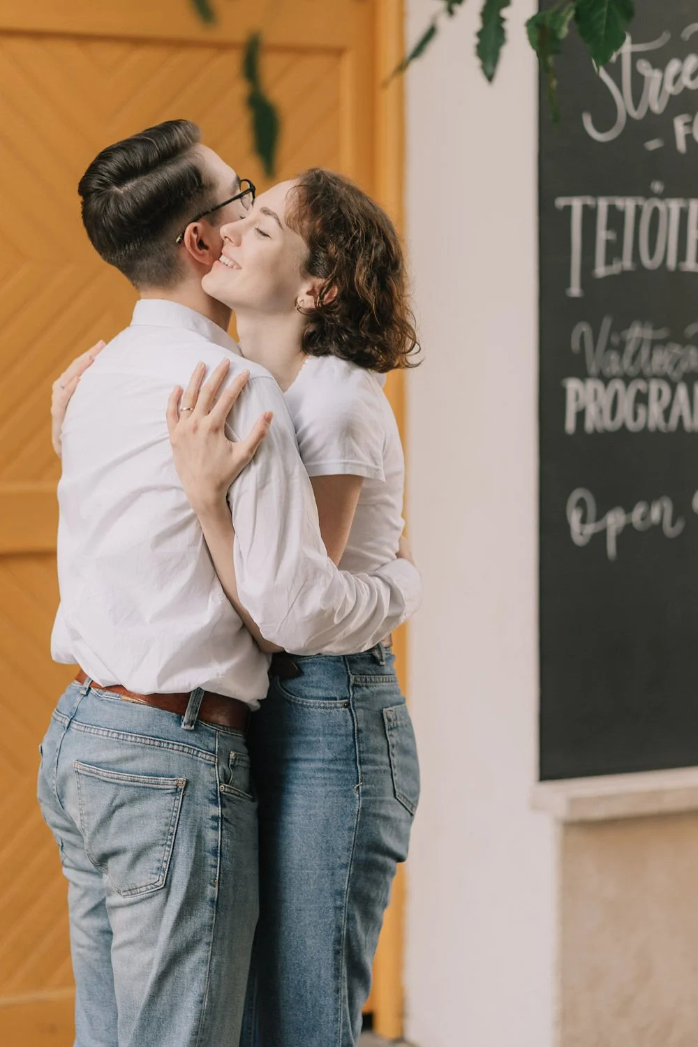 Casal se abraçando e sorrindo, usando roupas casuais, dentro de um ambiente interno com decoração de madeira ao fundo.