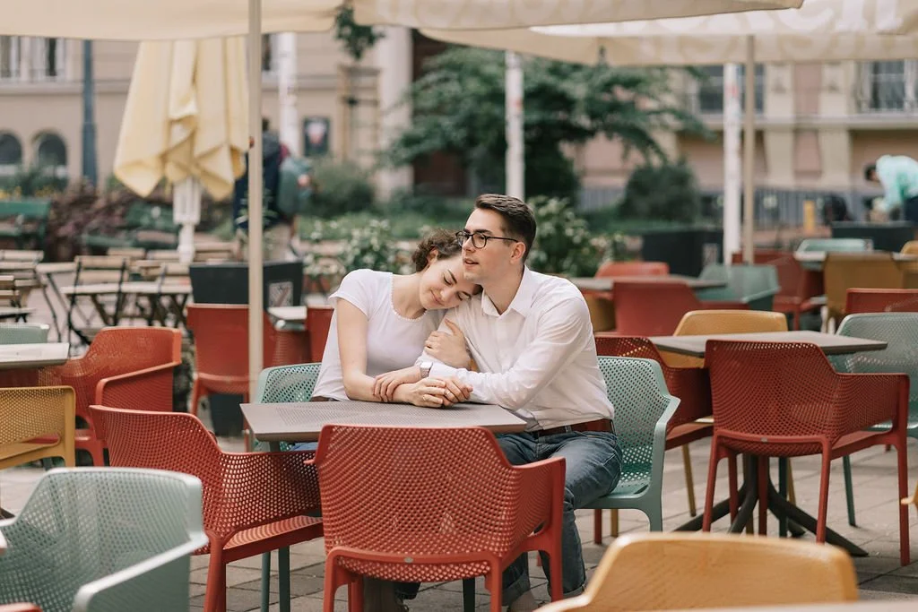 Casal de jovens sentados em mesa de café ao ar livre, com guarda-sol, em contexto urbano.
