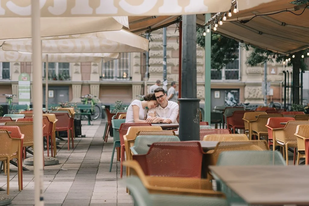 Casal de jovens sentados em uma mesa de um restaurante ao ar livre, com várias cadeiras coloridas e pérgolas com luzes decorativas.