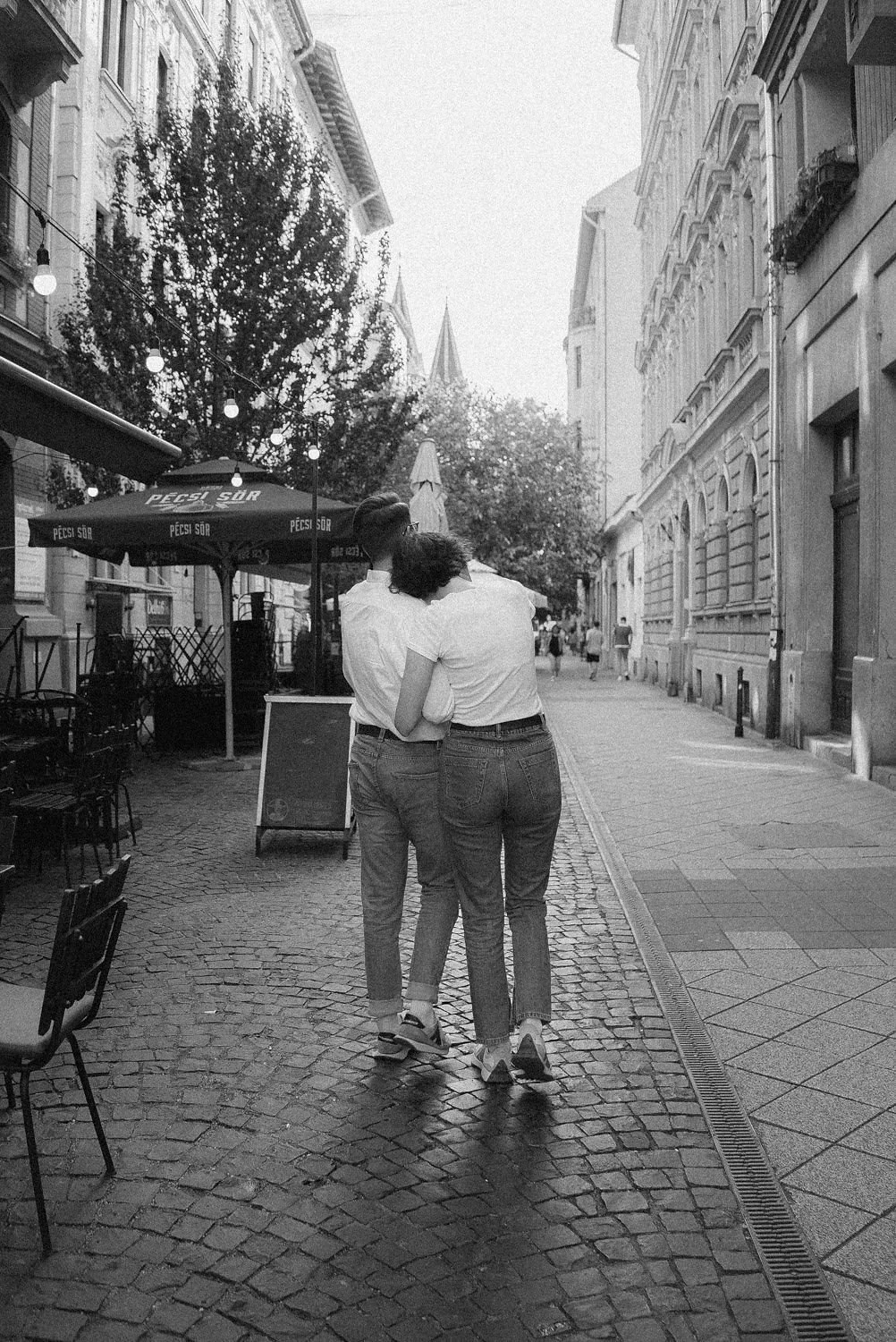 Casal caminhando de braços dados na rua de paralelepípedos, carsagens antigas e prédios históricos ao fundo, ao pôr do sol, em uma cidade.