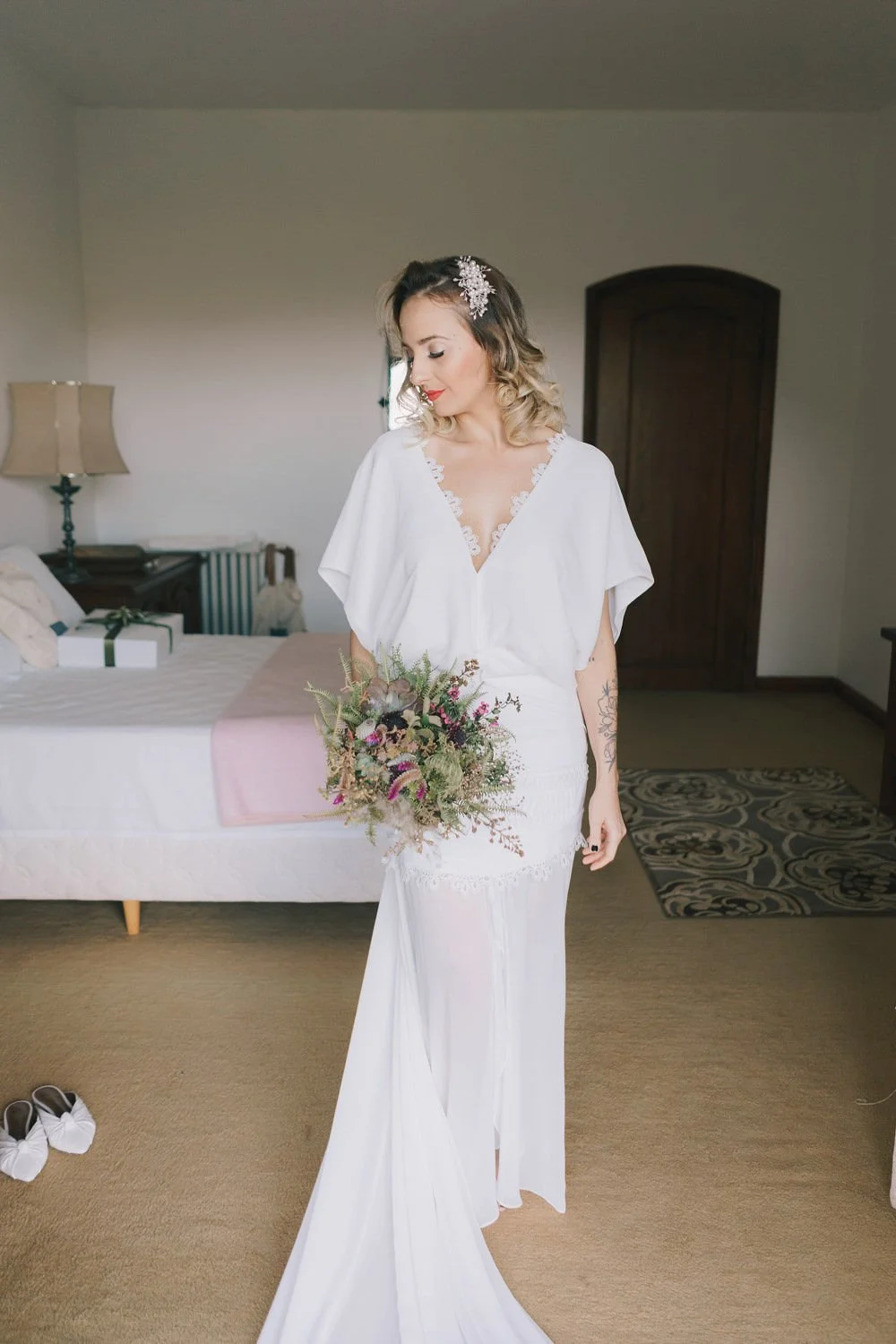 Mulher vestida de branco com vestido de noiva, segurando um buquê de flores, no interior de um quarto, com sapatos brancos no chão ao lado.