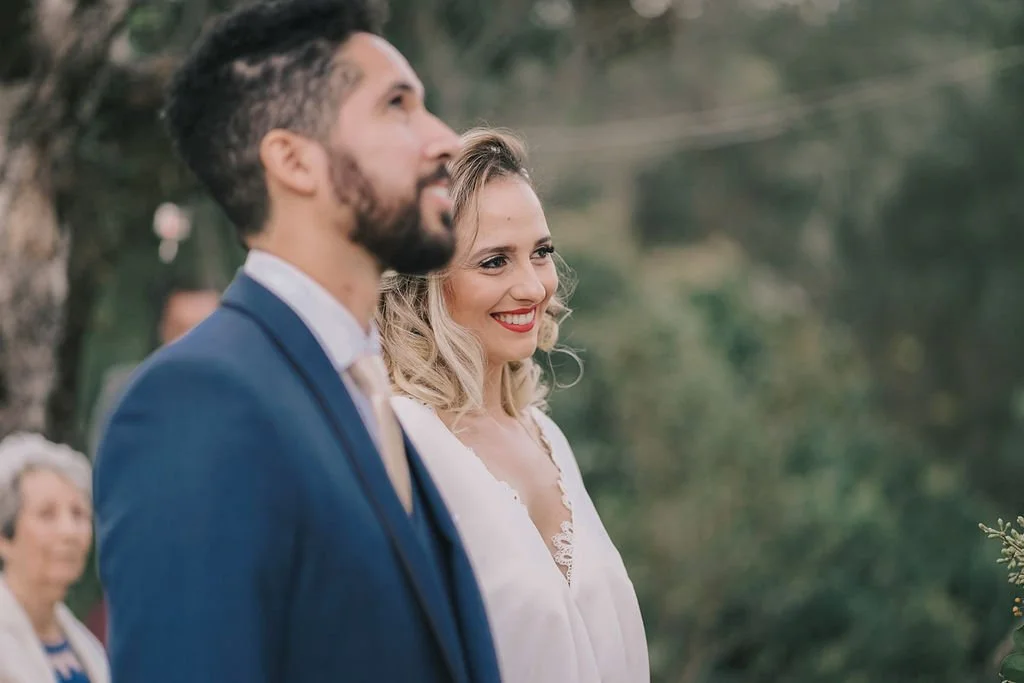Casal sorridente em cerimônia ao ar livre, com a mulher usando vestido branco e o homemja usando terno azul escuro.