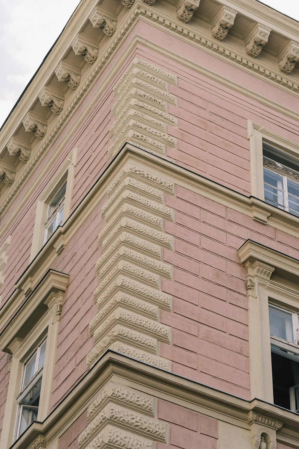 Imagem de uma esquina de um prédio antigo de cor rosa com detalhes em branco na fachada, incluindo janelas com moldura branca eornamentação decorativa acima das janelas.