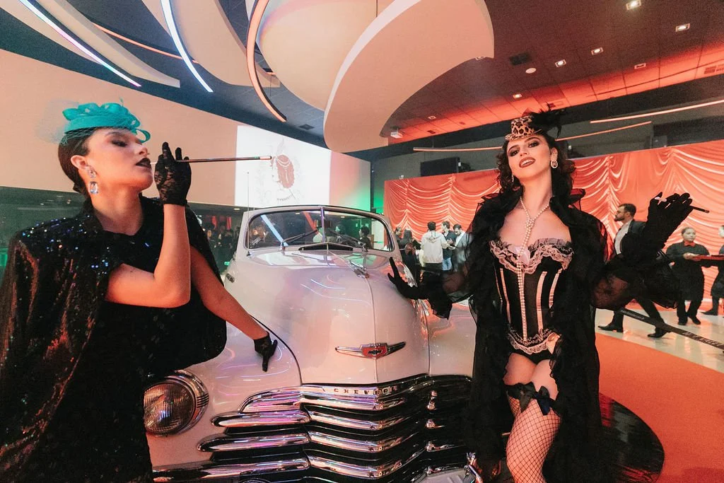 Mulheres vestidas com roupas elegantes e glamourosas posando ao lado de um carro clássico em um evento sofisticado. Uma delas fuma um cigarro de tonalidade escura e usa chapéu azul, enquanto a outra usa roupa de lingerie com uma capa preta e luvas.