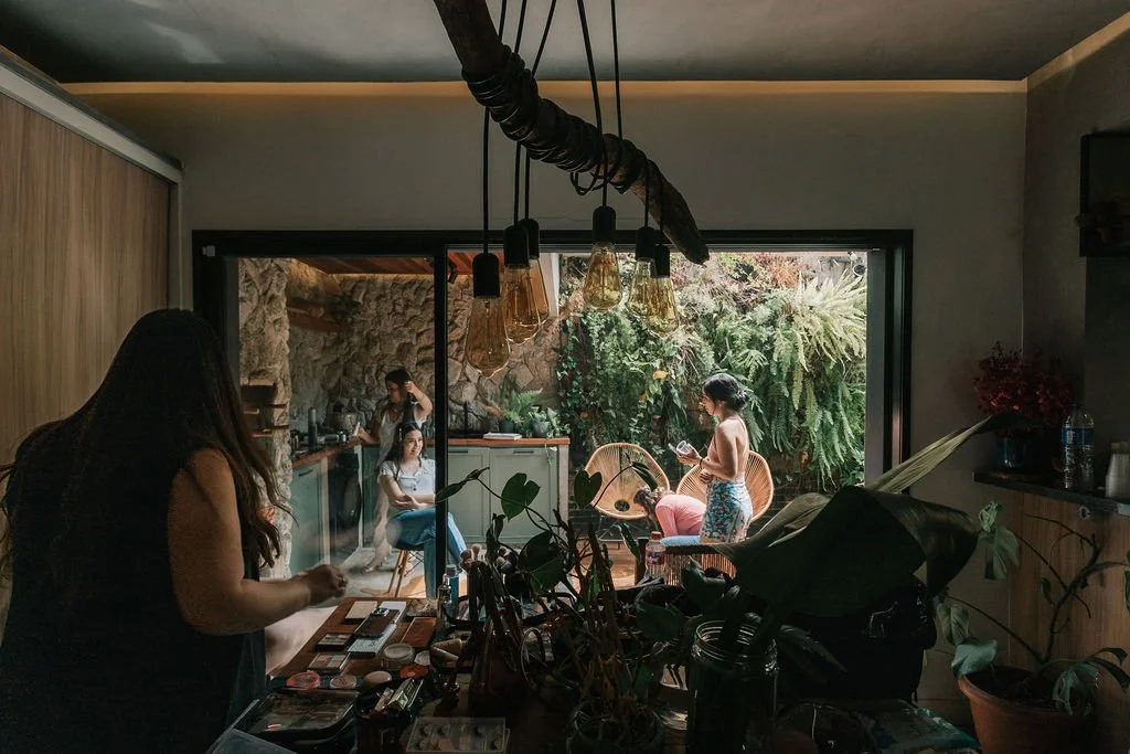 Interior de uma casa com pessoas na varanda. Uma mulher de cabelo cacheado e liso de costas, na sala, com objetos e plantas na mesa. Na varanda, há uma mulher com camiseta cinza, uma mulher de shorts cor de rosa e uma outra em uma cadeira com celular