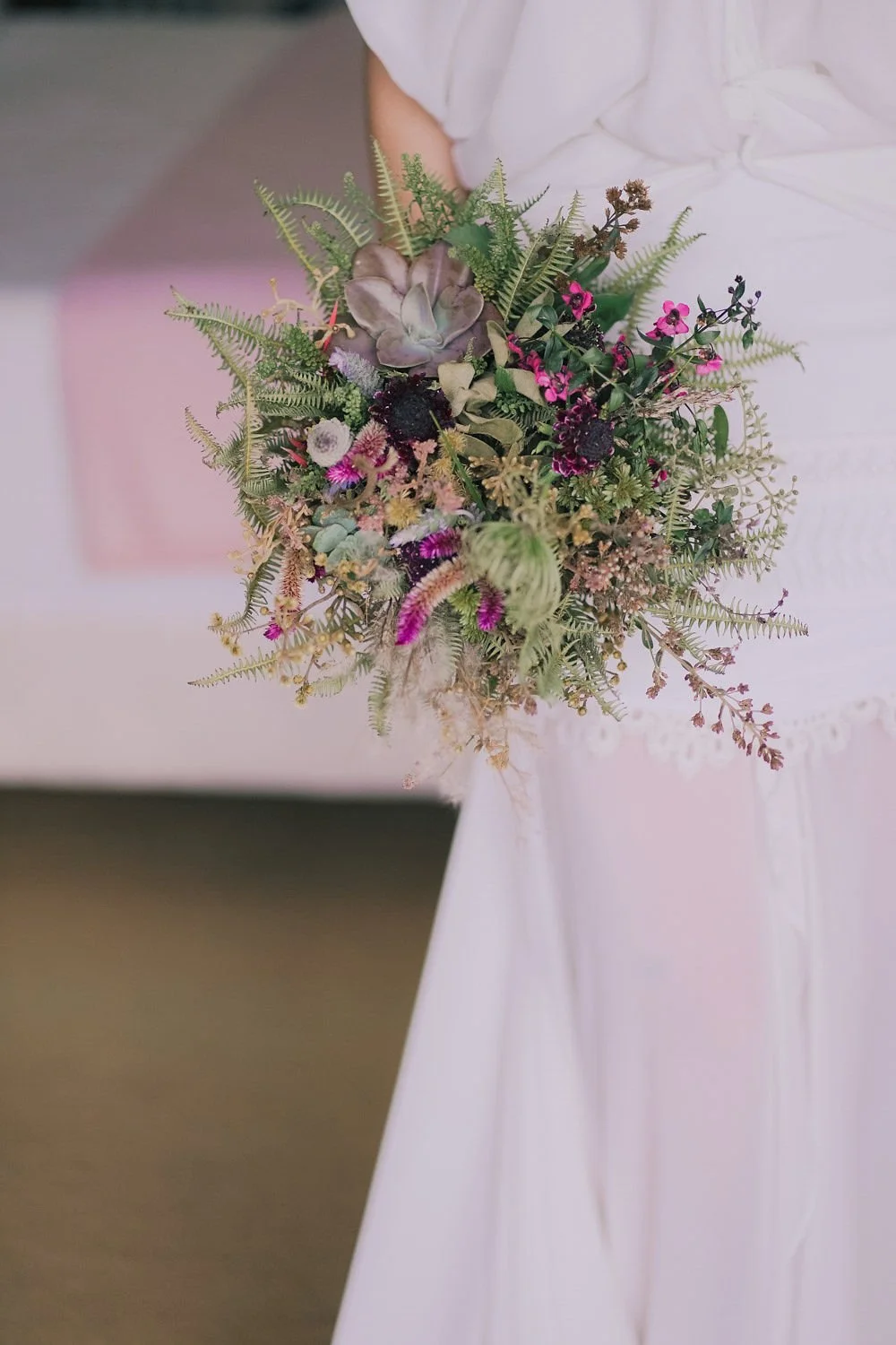 Primeiro plano de um buquê de flores com várias plantas e flores coloridas e verdes, sendo segurado por uma pessoa com vestido branco.