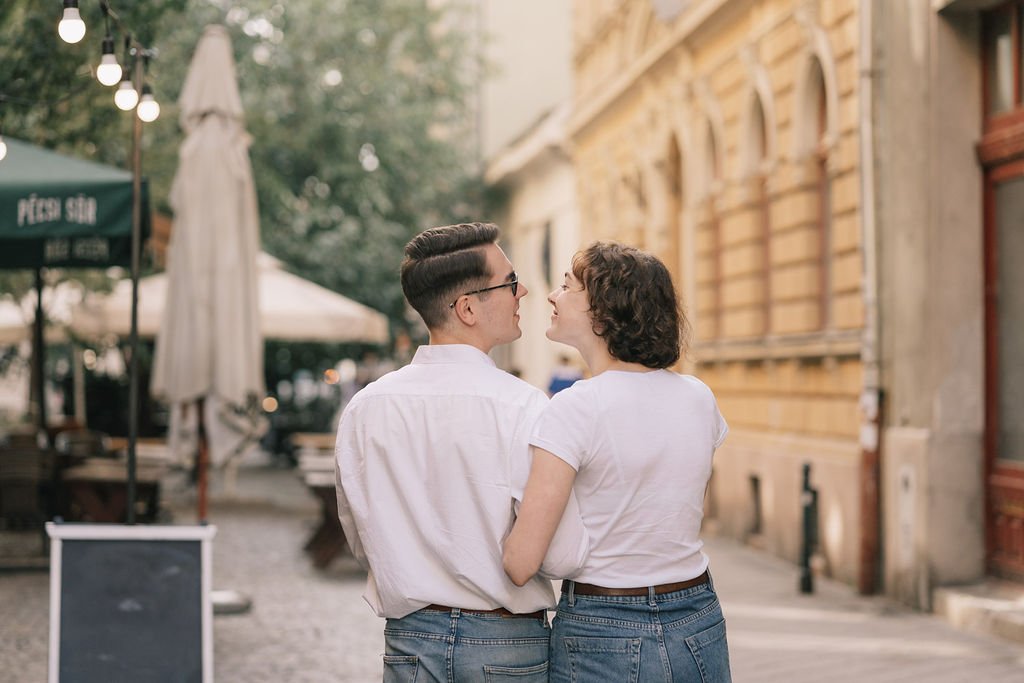 Casal caminhando e conversando na rua de paralelepípedos, com decoração de luzes, ao lado de um edifício antigo, sob um céu claro.