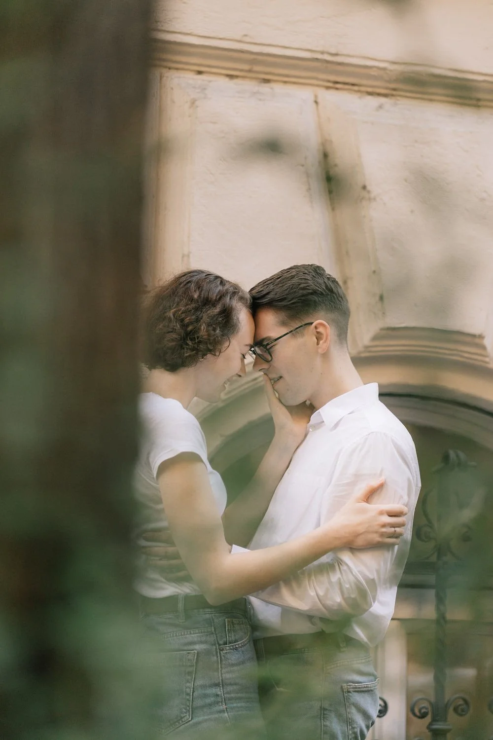 Imagem de um casal jovem, de cabelos castanhos e pele clara, tocando a testa e sorrindo, com a cabeça empenhada uma na outra, em um espaço com paredes de pedra e ferro decorativo ao fundo.