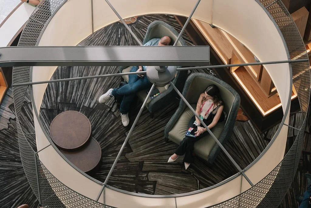 Vista de cima de duas pessoas sentadas em poltronas dentro de uma estrutura circular com iluminação de LED, com duas mesas pequenas entre elas, em um espaço de leitura ou espera.