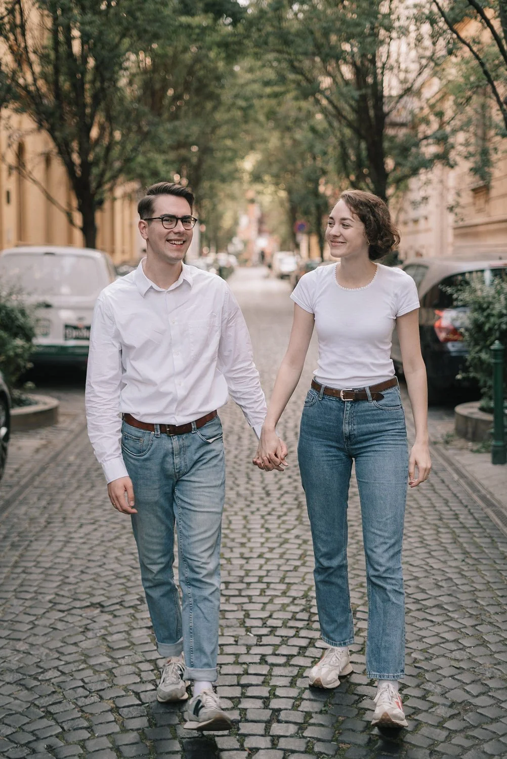 Casal caminhando de mãos dadas na rua de paralelepípedo, cercados por árvores e carros estacionados, durante o dia.