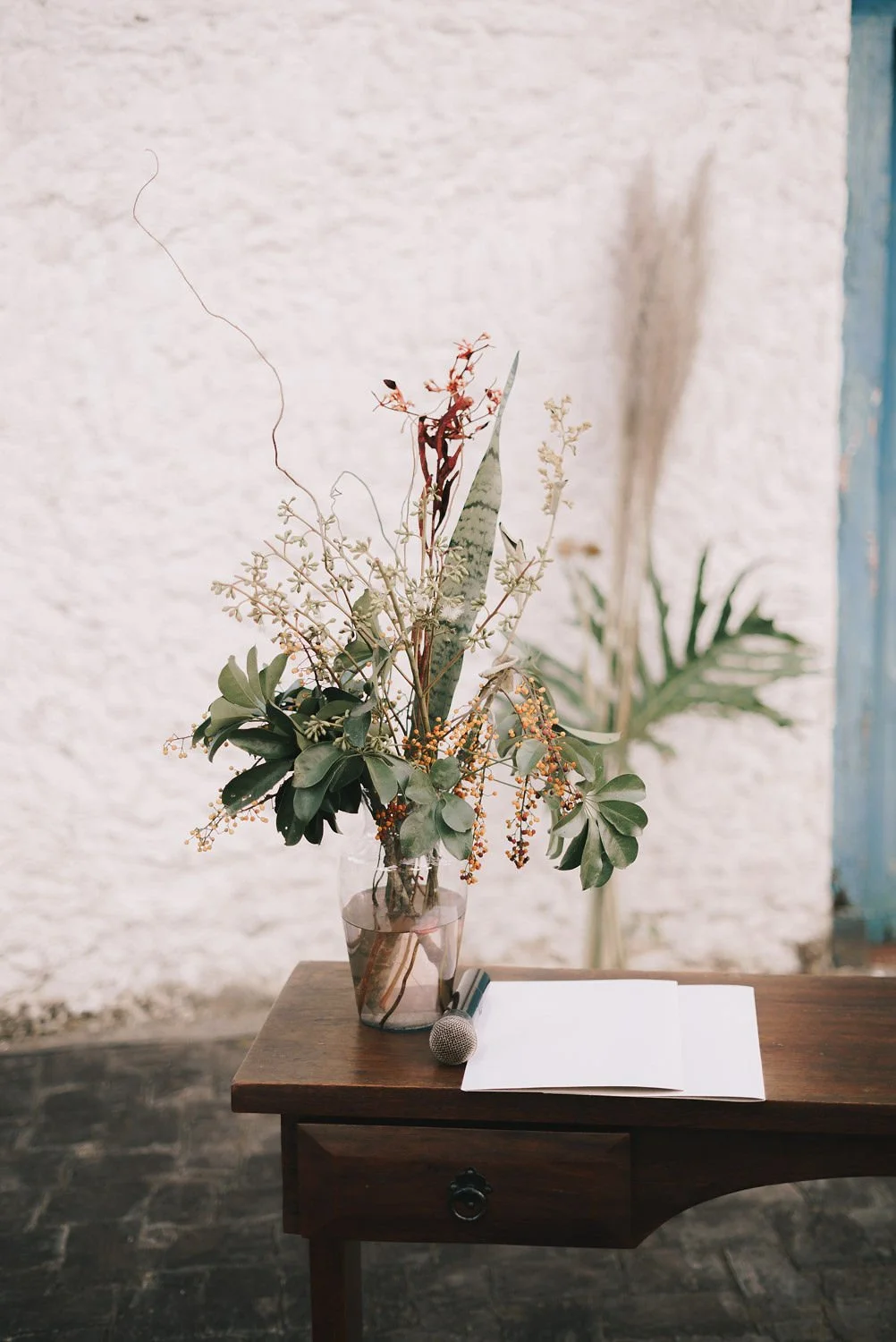 Arranjo de flores com folhas verdes variadas em um vaso de vidro sobre uma mesa de madeira, ao lado de um microfone e um livro aberto.