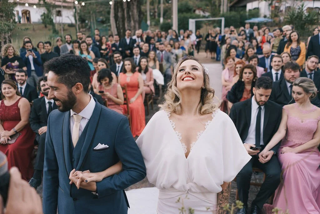 Noivos em cerimônia de casamento ao ar livre, com muitas pessoas na plateia sorrindo ao fundo.
