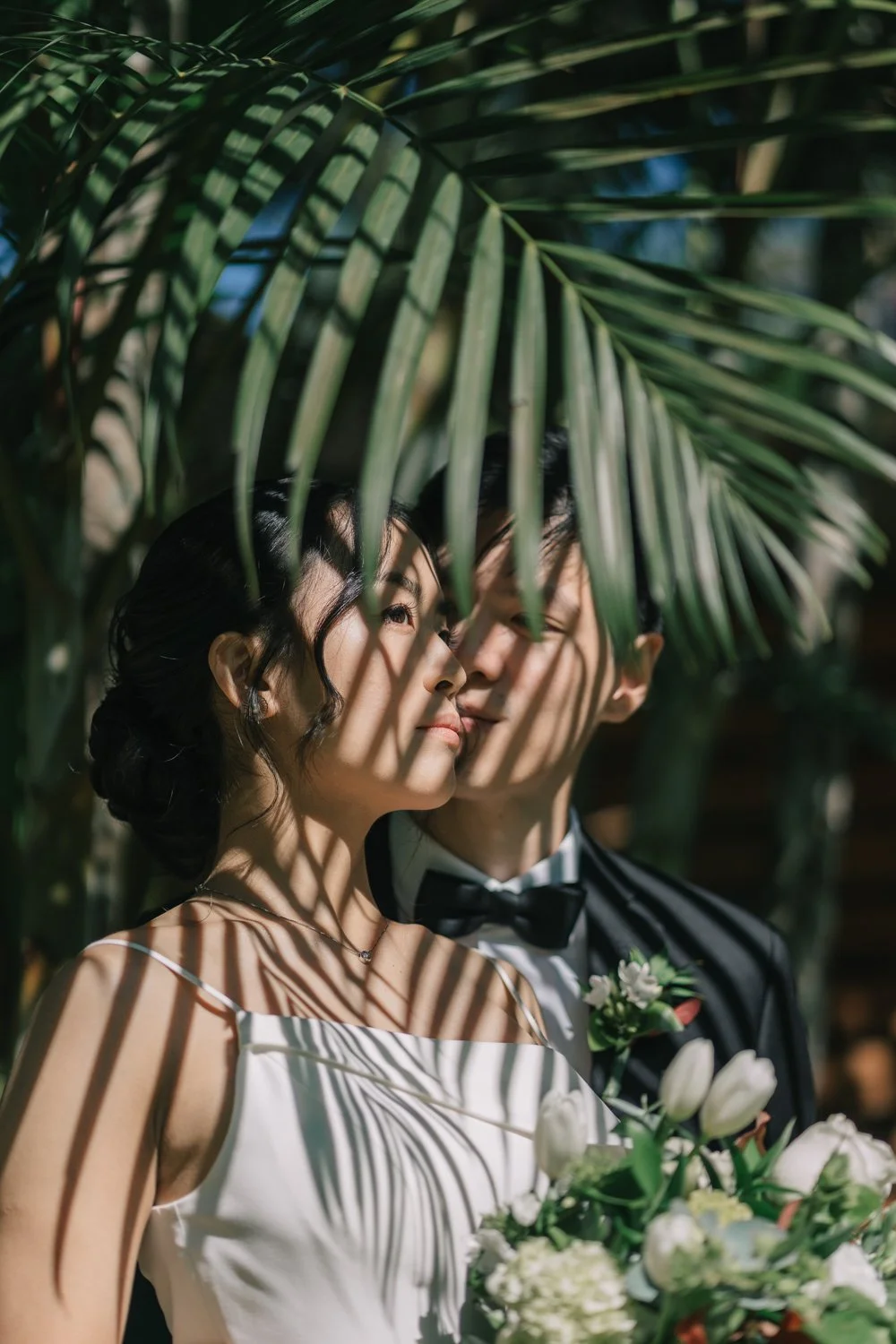 Casal de noivos em casamento sob a sombra de uma folha de palmeira, com luz filtrada criando sombreamento e destaque no rosto dos noivos, segurando flores brancas.