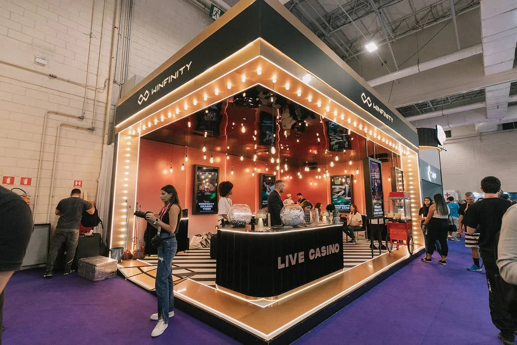 Espaço de um evento com estande de cassino ao vivo da Winfinity, decorado com luzes pendentes e iluminação de energia quente, com pessoas interagindo e desfrutando da atividade.