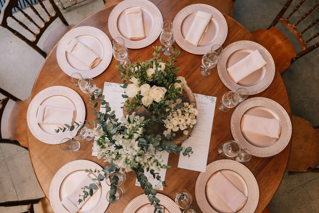 Mesa de jantar decorada com flores brancas e copos de vidro, com pratos e talheres brilhantes, vista de cima.