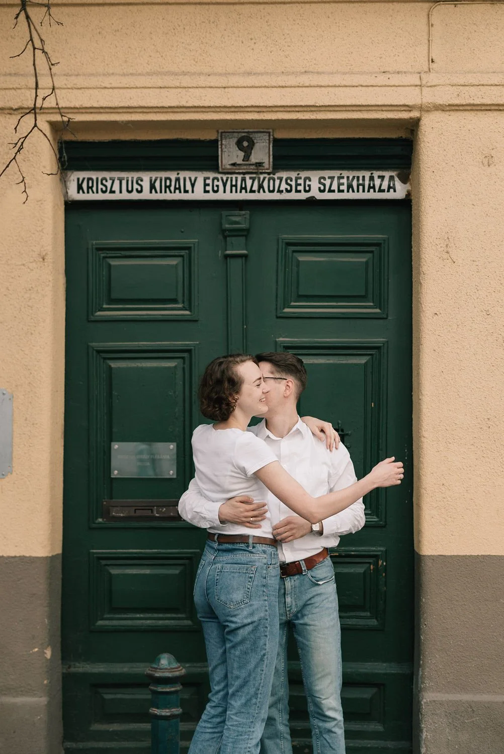 Um casal se abraçando e se beijando na rua, na frente de uma porta verde, com sinalização em húngaro. Eles parecem felizes e afetuosos.