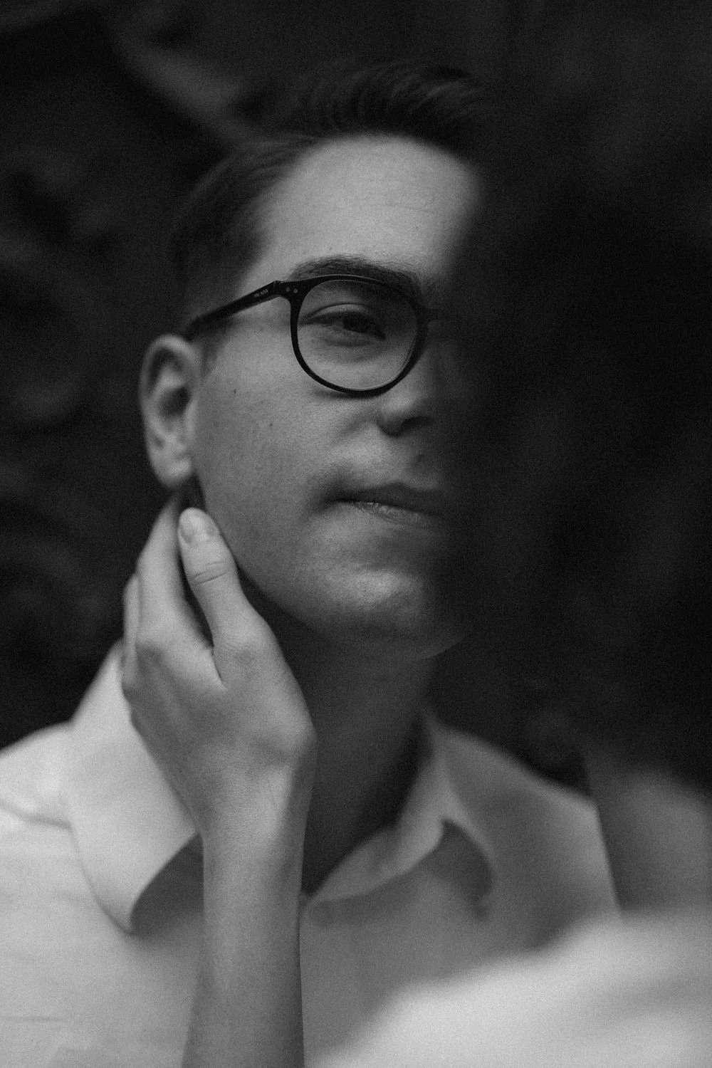 Retrato preto e branco de uma pessoa usando óculos, com expressão serena, tocando o pescoço com uma mão, em fundo escuro.