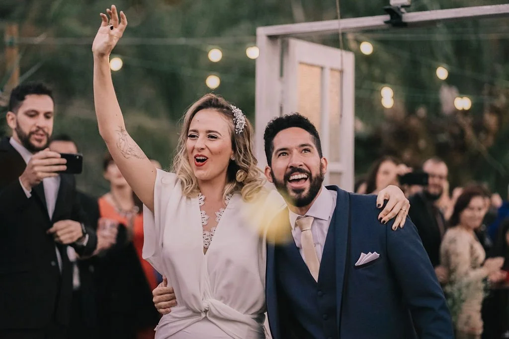 Casal sorridente em celebração ao ar livre na noite, rodeado de convidados, com iluminação decorativa.