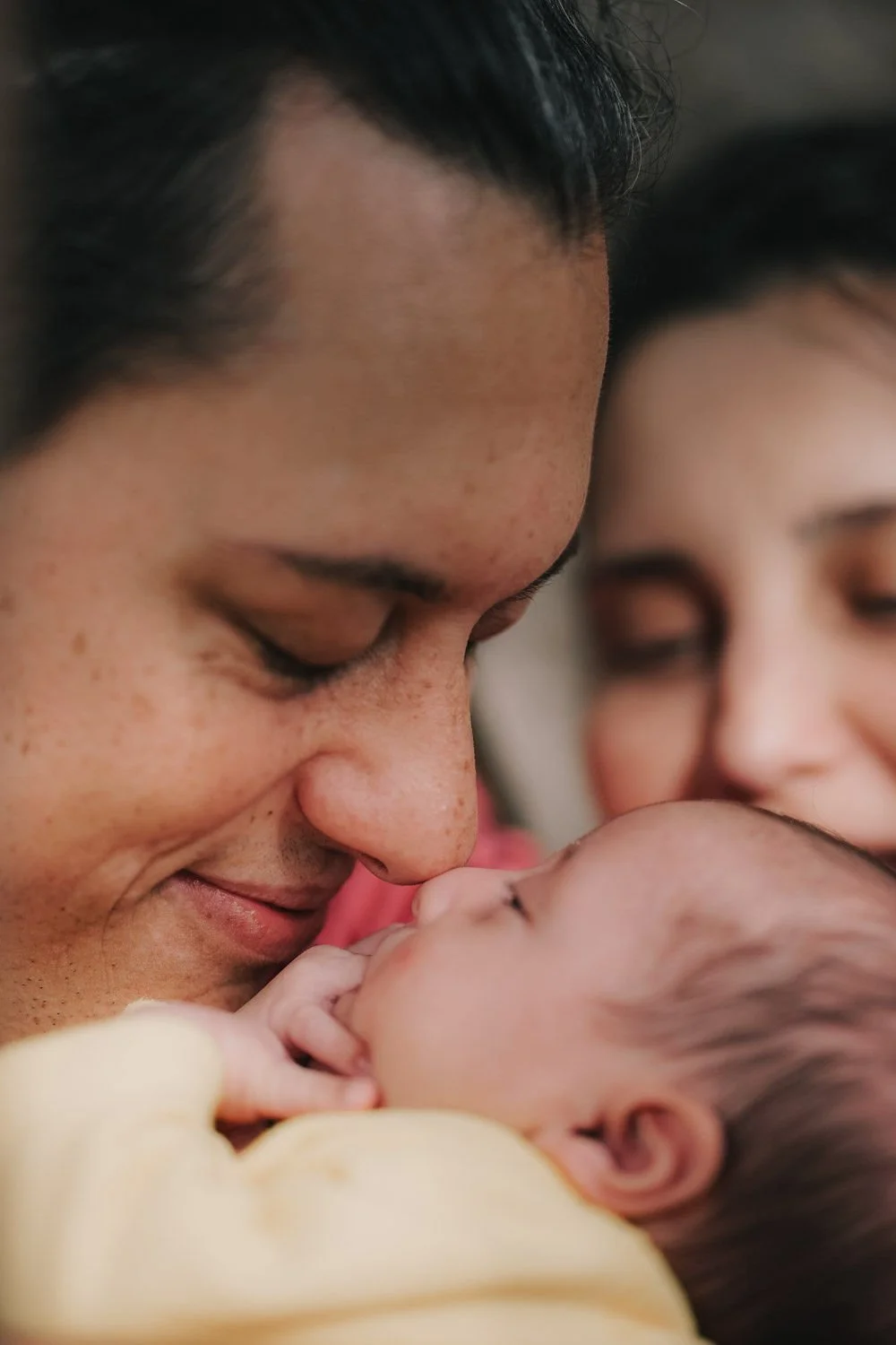 Imagem de uma pessoa sorrindo enquanto segura um bebê, com outra pessoa ao fundo.