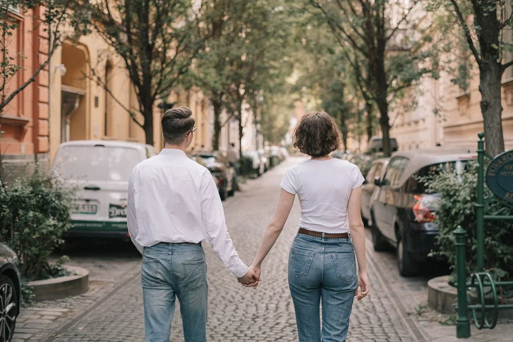 Casal andando de mãos dadas na rua com árvores e carros estacionados.